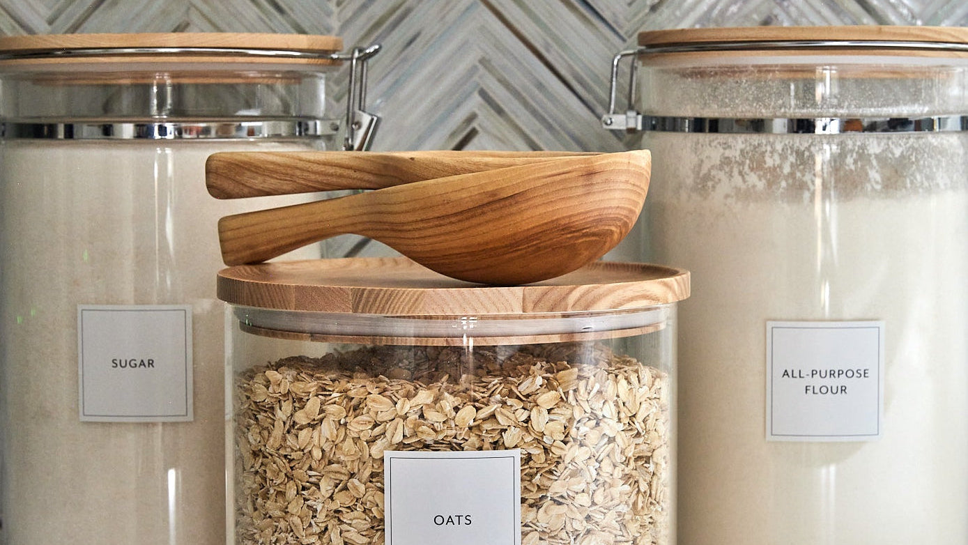 Three glass storage jars with wooden lids on a kitchen counter, labeled for different pantry items.